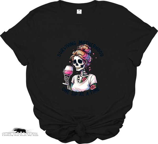 Black t-shirt with a colorful graphic of a skeleton holding a cocktail and text 'Surviving Motherhood One Sip at a Time' on a white background.