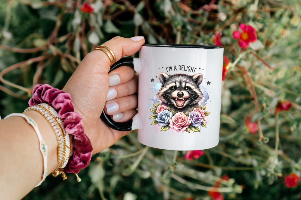 Hand holding a mug with a raccoon illustration and floral design, against a natural background.
