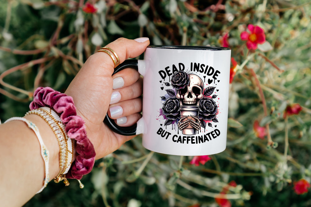 Hand holding a mug with a skull and roses design, text 'Dead Inside But Caffeinated', against a natural background.