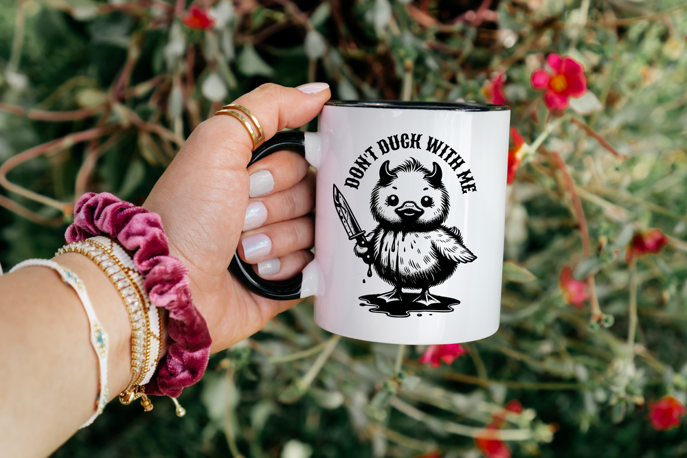 Hand holding a mug with a graphic design of a duck holding a sword and text 'Don't Duck with Me' against a natural background.