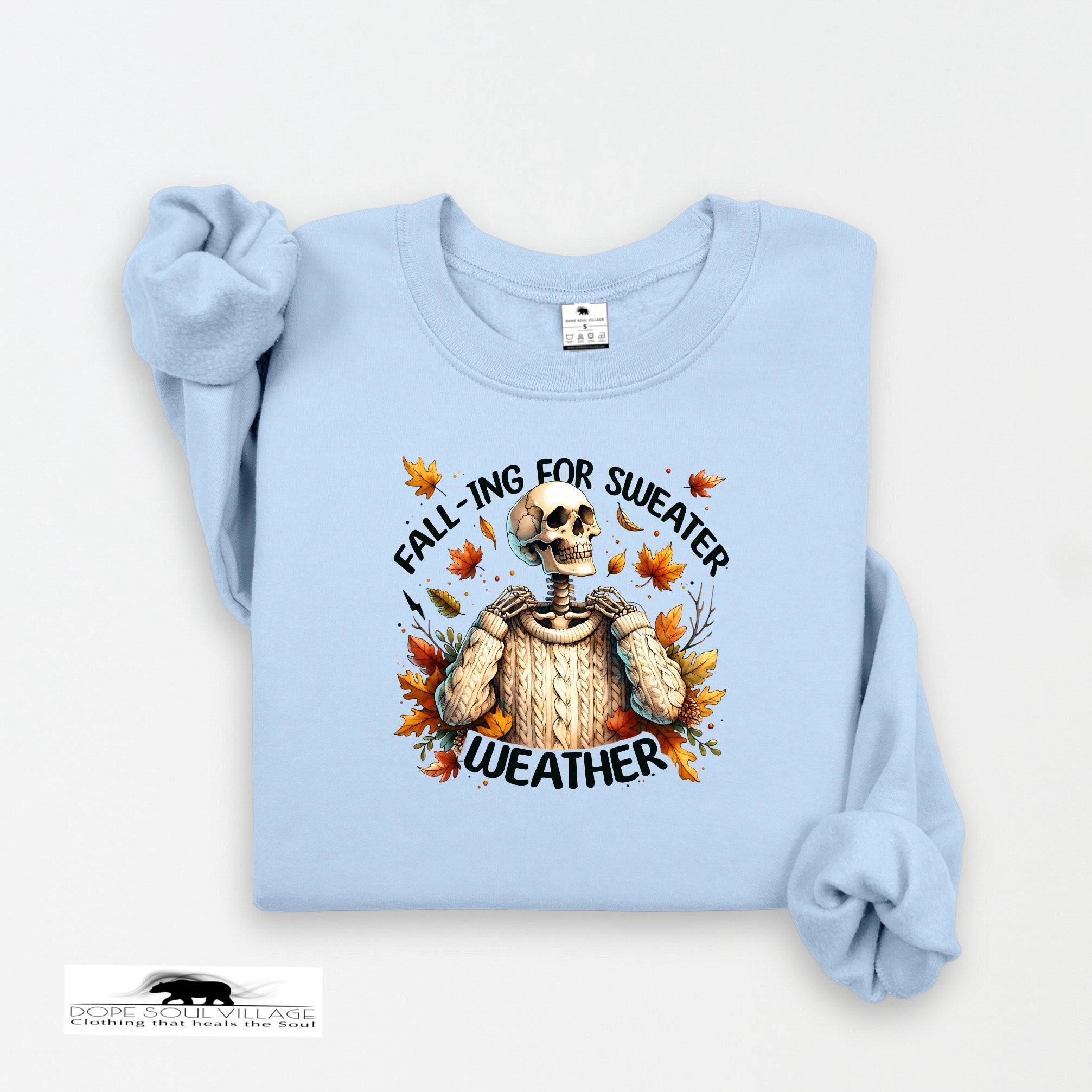 Light blue sweatshirt with skeleton wearing a sweater and autumn leaves design on a white background.