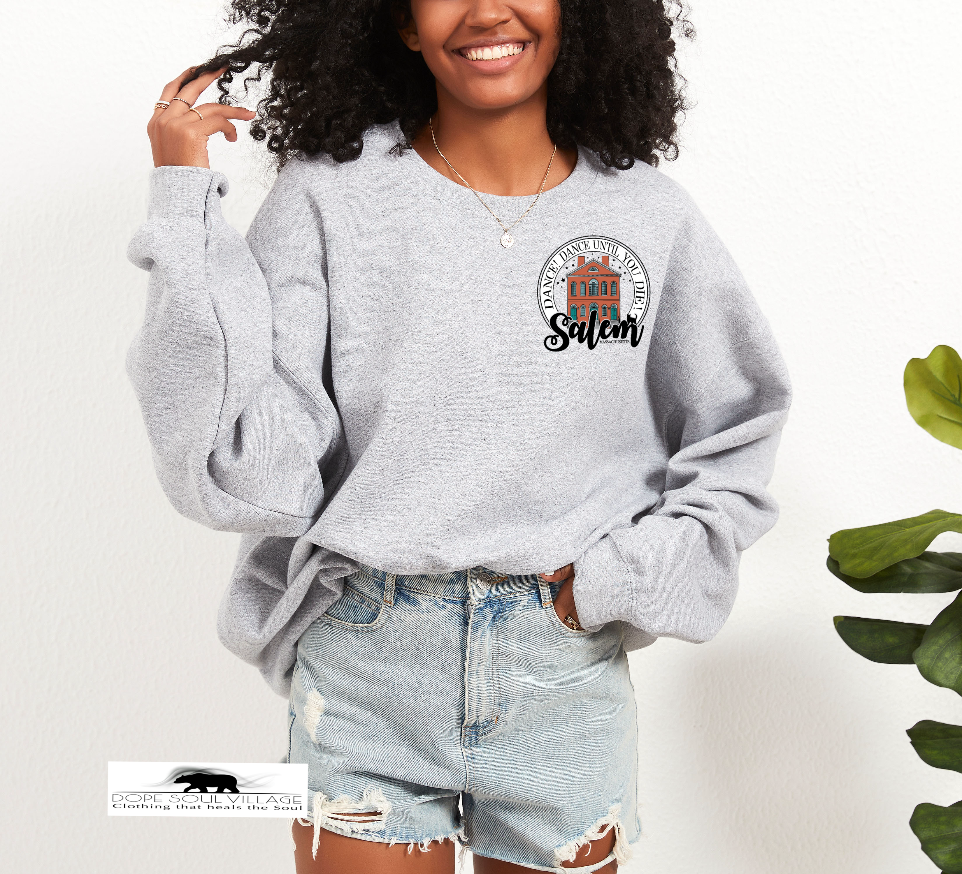 Woman wearing a gray sweatshirt with a logo and text, standing against a white background salem sister halloween collection dope soul village
