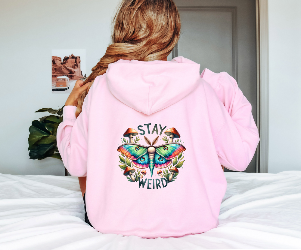 Person wearing a pink hoodie with a colorful butterfly design and 'Stay Weird' text on the back, sitting on a bed.