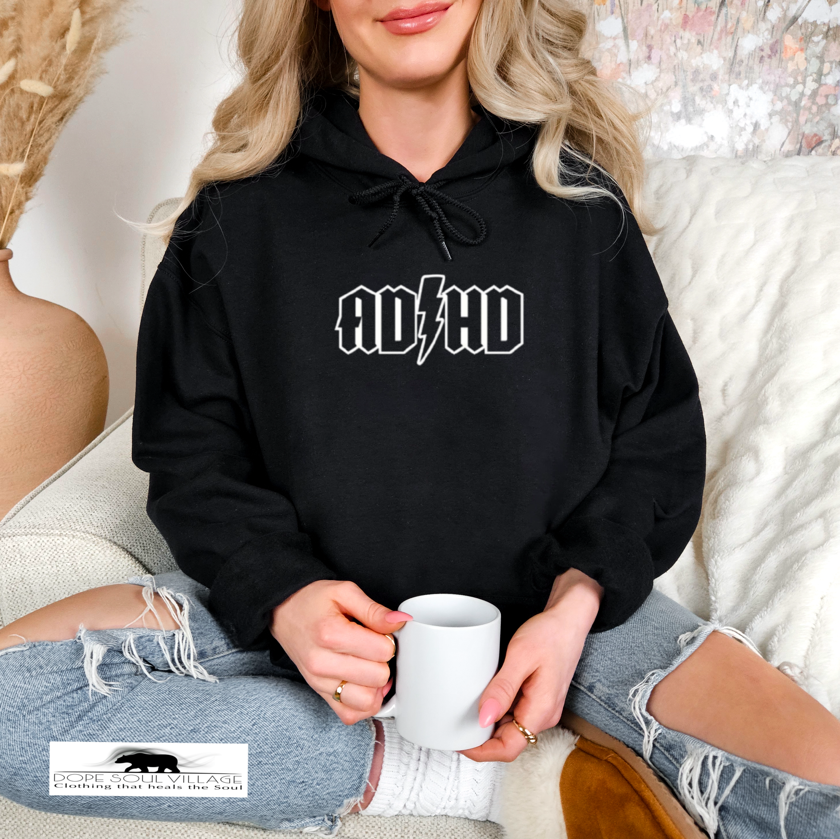 Person wearing a black hoodie with 'ADHD' text, holding a white mug, sitting on a couch.