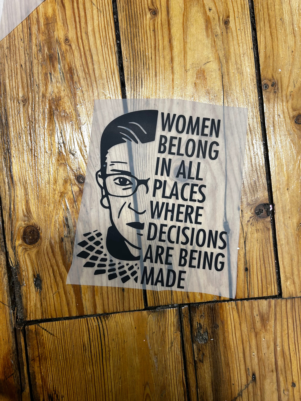 A square printed sticker or decal with black text and a stylized face illustration, placed on a rustic wooden plank surface. The text reads, 'WOMEN DECISIONS ARE BEING MADE WHERE PLACES BELONG IN ALL'.
