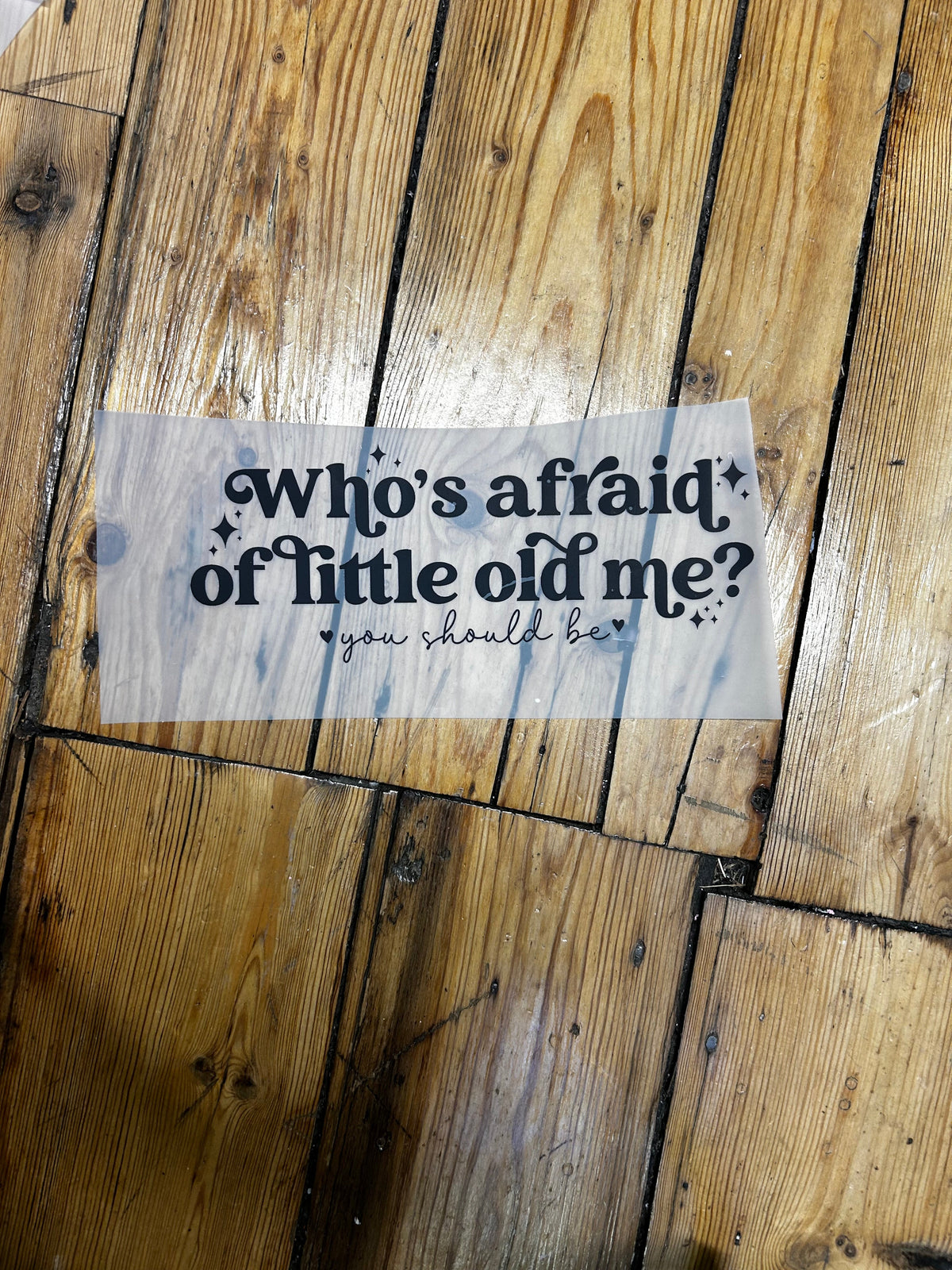 A rectangular stencil with black text reading 'Who's afraid of little old me?' and small star accents, placed on a rustic wooden plank floor.