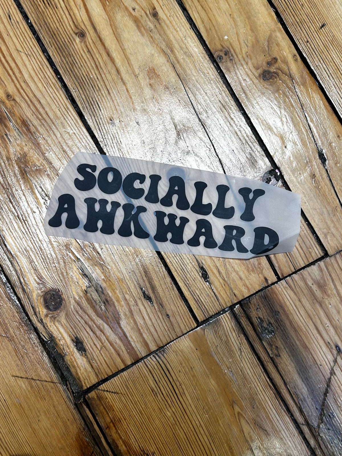 A die-cut sticker with the word 'ATTACK' in black letters on a translucent white backing, placed on a rustic wooden floor.