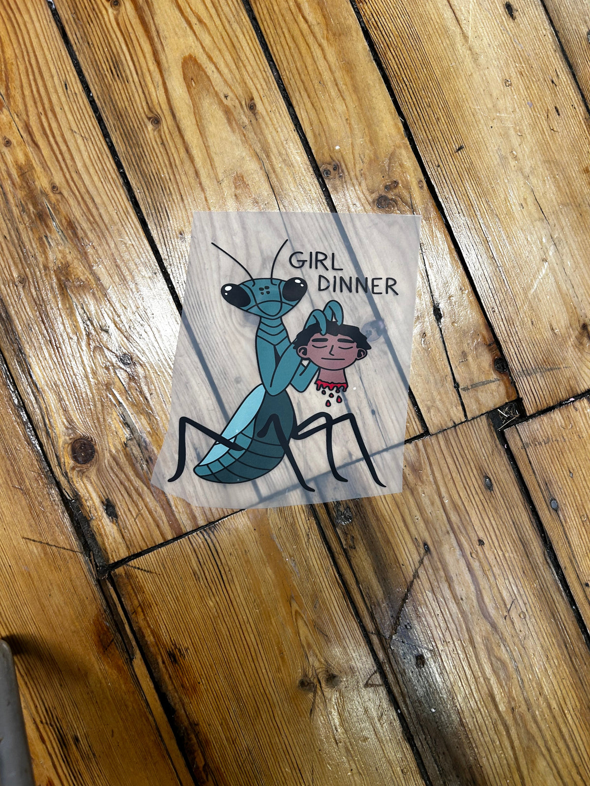 A square clear sticker with a cartoon graphic of a girl in a blue swimsuit holding a pink drink, placed upside down on a wooden floor. The sticker has the words 'GIRL DINNER' printed upside down in black.