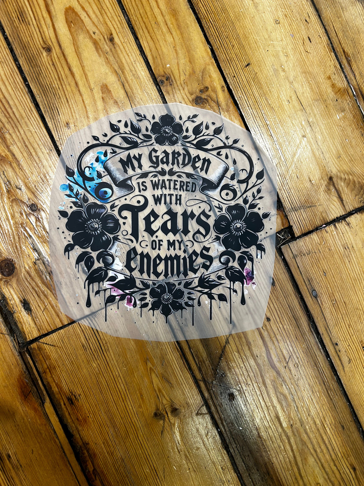 A circular clear vinyl sticker with black gothic-style text and floral illustrations, placed on a rustic wooden plank floor.