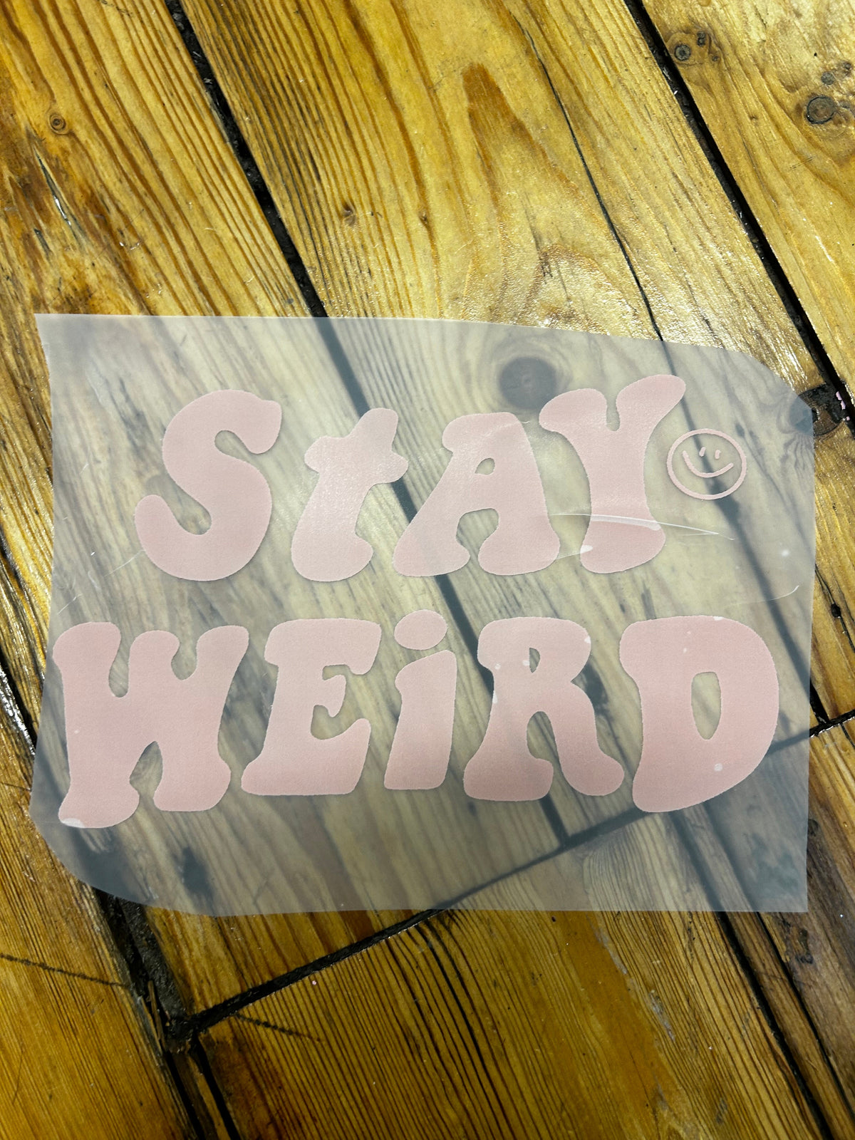 A pink vinyl decal sticker with the text 'STAY WEIRD' in a retro bubble font, shown on a translucent backing sheet over a wooden plank floor.