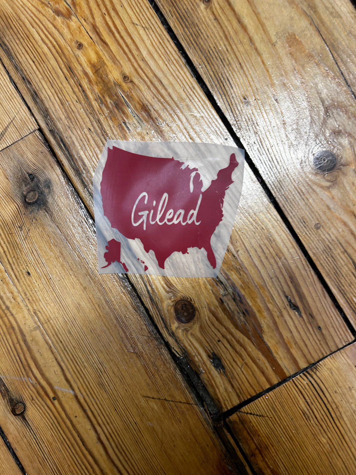 A square sticker with a burgundy silhouette of Wales and the word 'Gilead' in white script, placed on a rustic wooden floor.