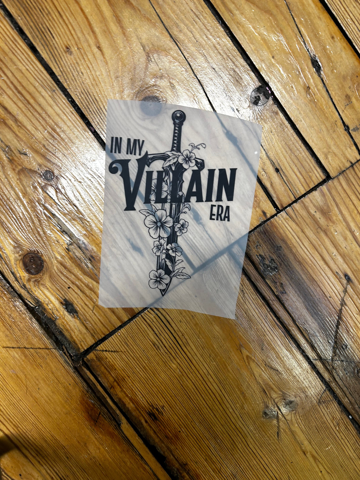 A rectangular printed sticker or decal with a black sword-and-floral graphic and the text 'VILLAIN ERA IN MY' placed on a wooden floor.