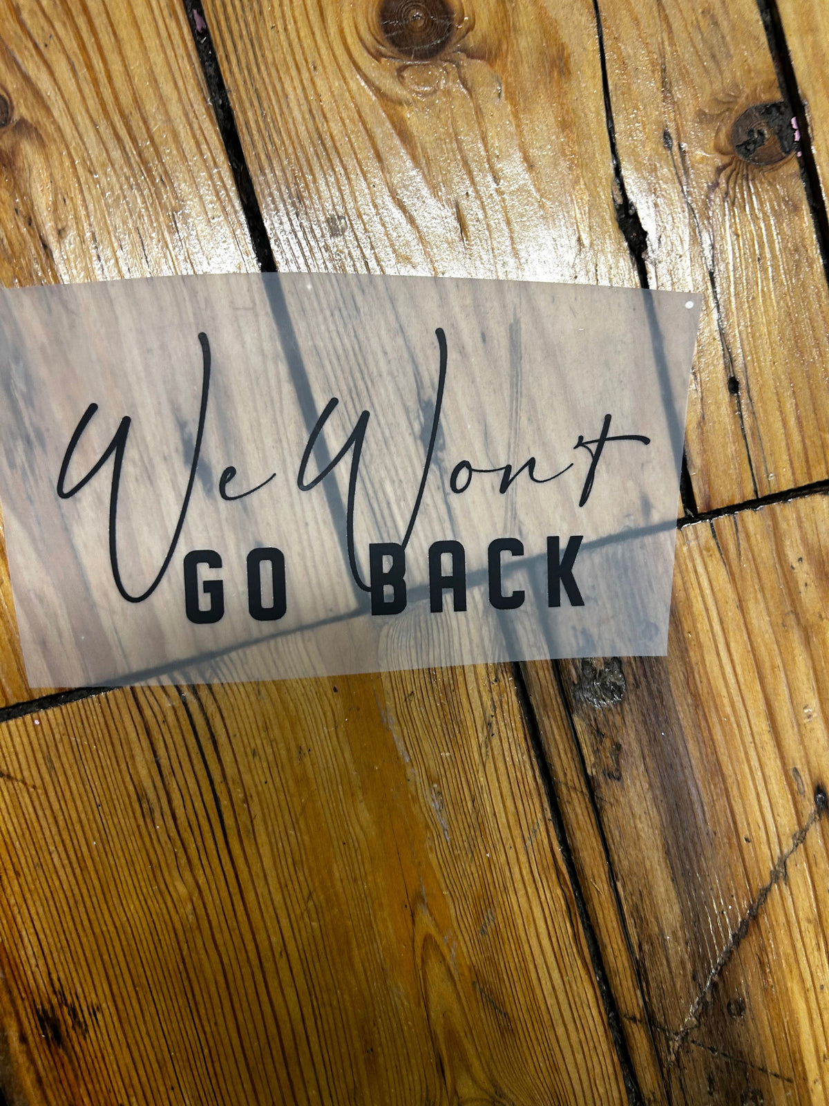 A vinyl decal sticker with the text 'GO BACK' and 'WELCOME' in black, shown on a translucent carrier sheet over a rustic wooden plank floor.