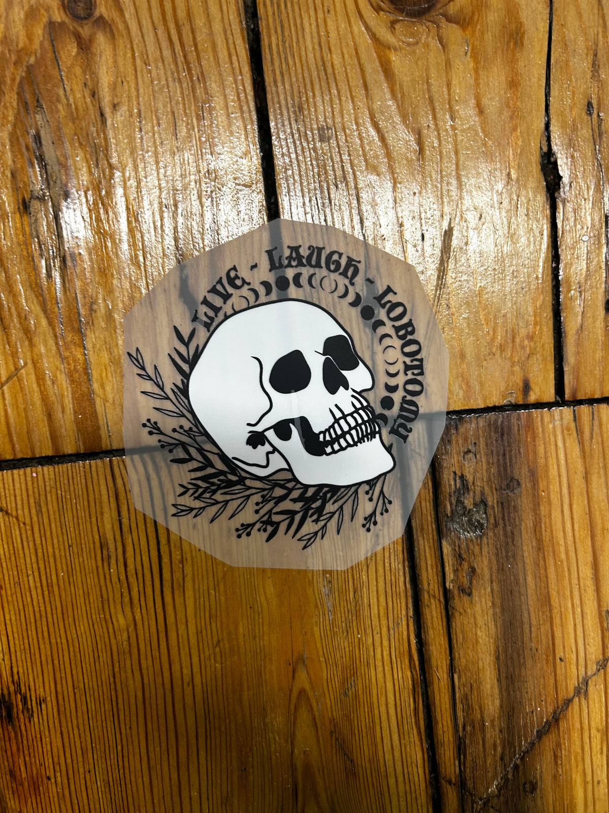 A circular die-cut sticker featuring a white skull with black eye sockets and black decorative leafy branches, placed on a glossy wooden plank surface. The sticker text reads 'LORPOE' and 'LACER' around a central design.