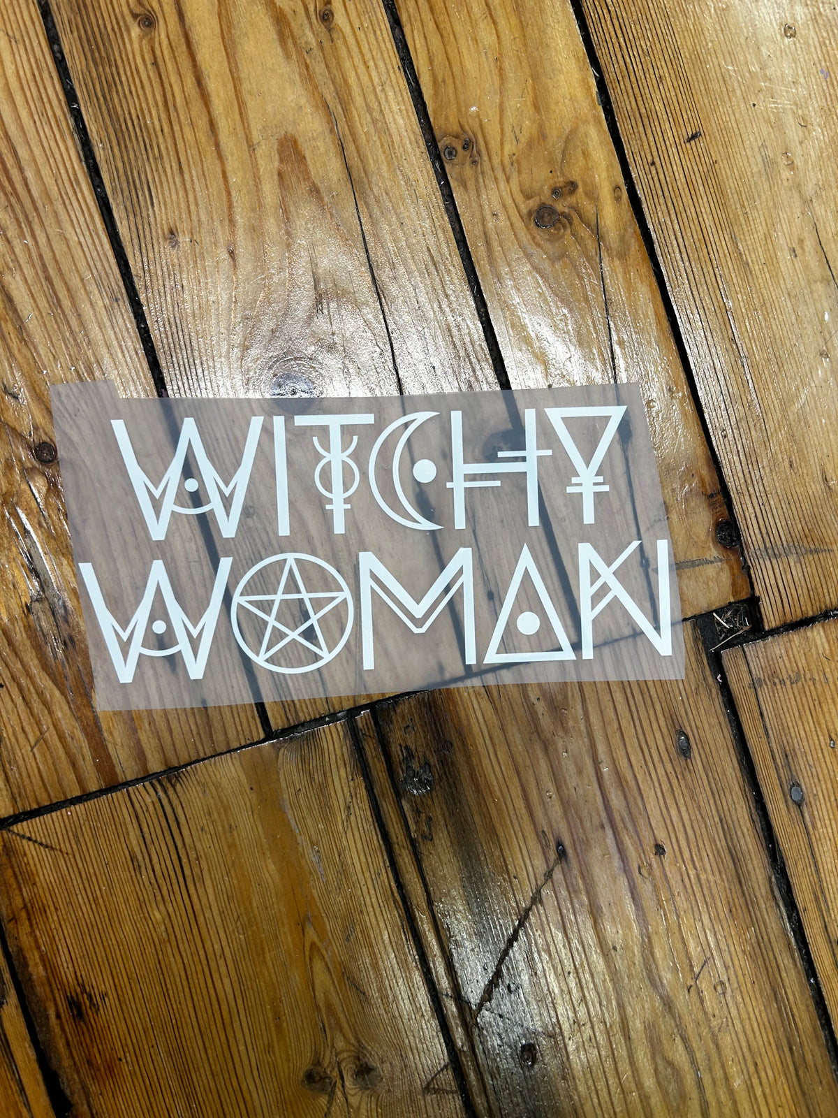 A rectangular clear vinyl decal sheet featuring white occult and witch-themed symbols, including a pentagram, moon, and stylized text, placed on a rustic wooden plank floor.