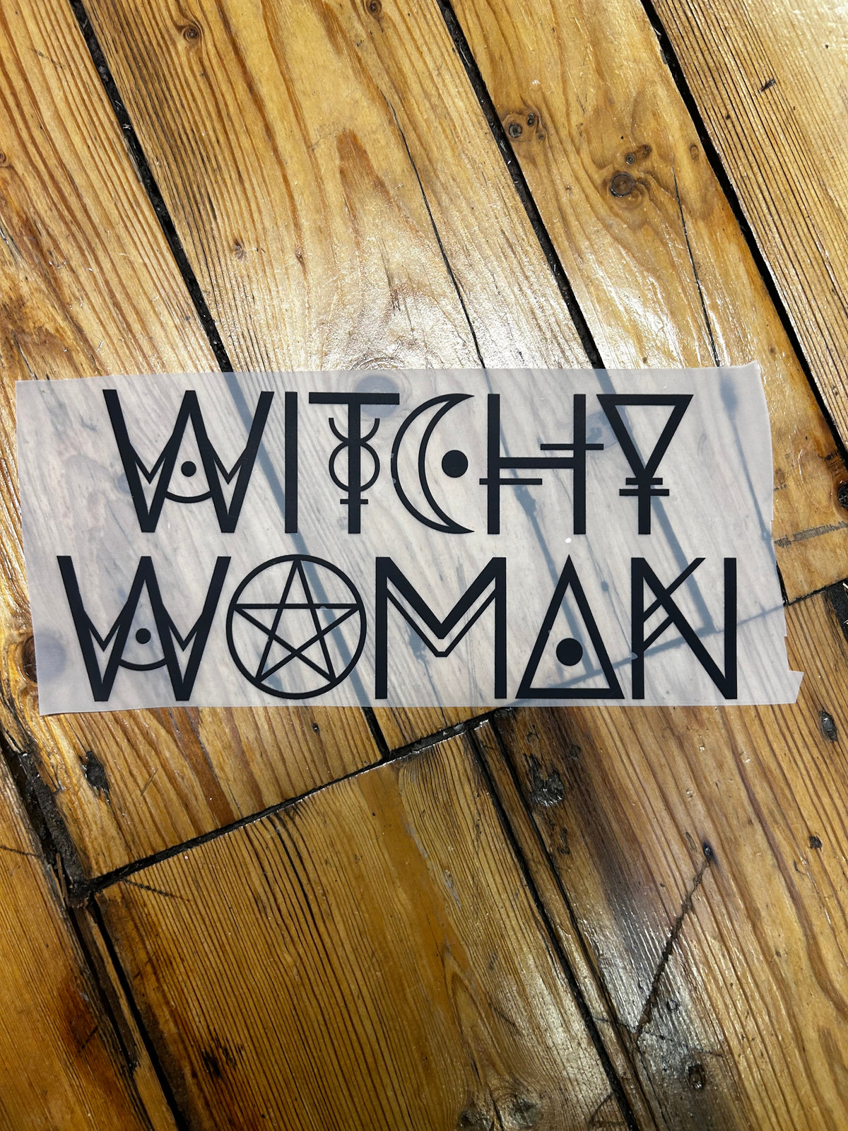 A vertical rectangular stencil or decal sheet with black occult-style lettering and symbols, placed on a rustic wooden plank floor.