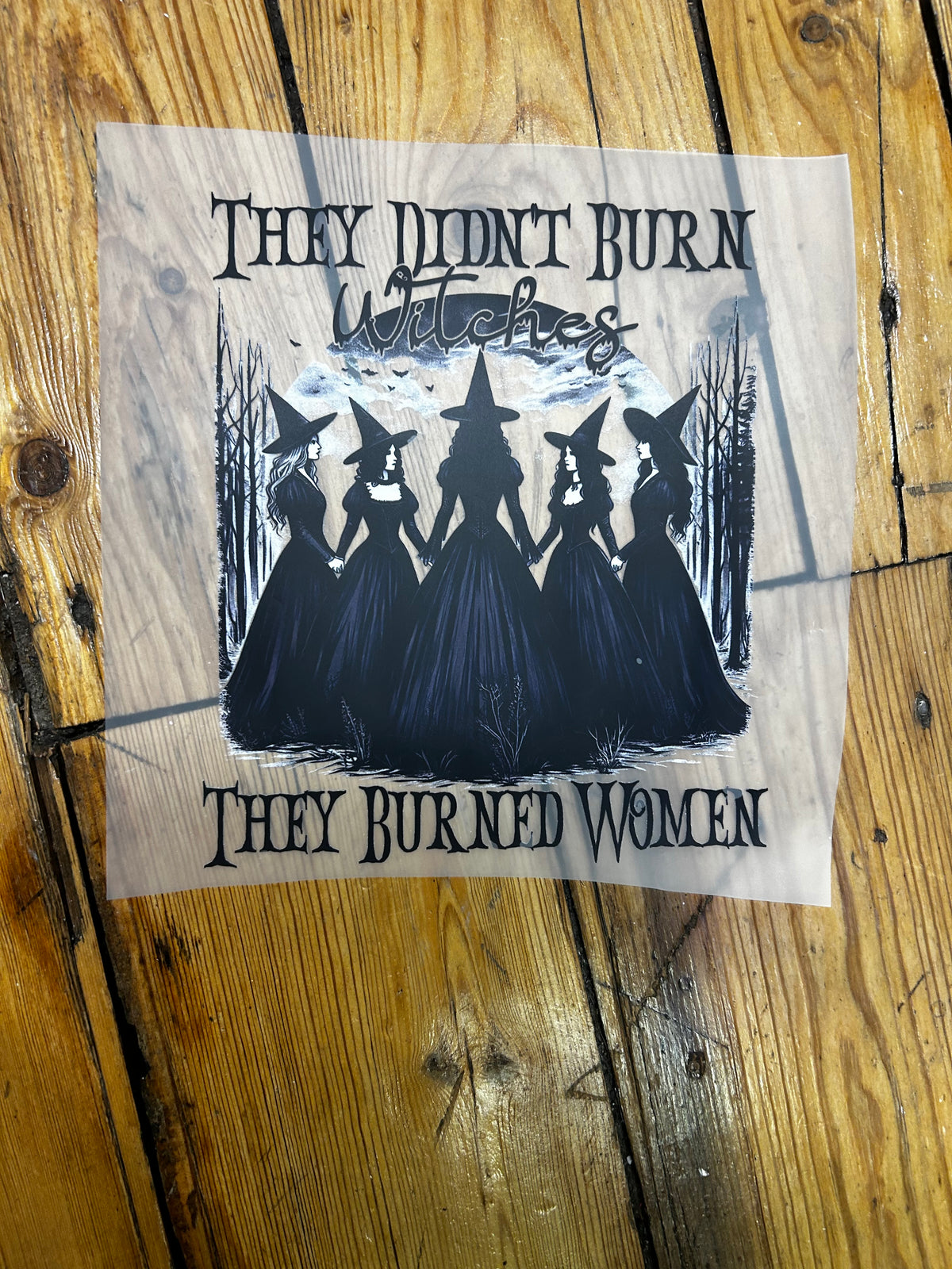 A square printed graphic on a piece of white paper, placed on a rustic wooden surface. The design shows five witch silhouettes in black dresses and hats under a pale sky, with the text 'THEY DIDN'T BURN Witches' at the top and 'THEY BURNED WOMEN' at the bottom.