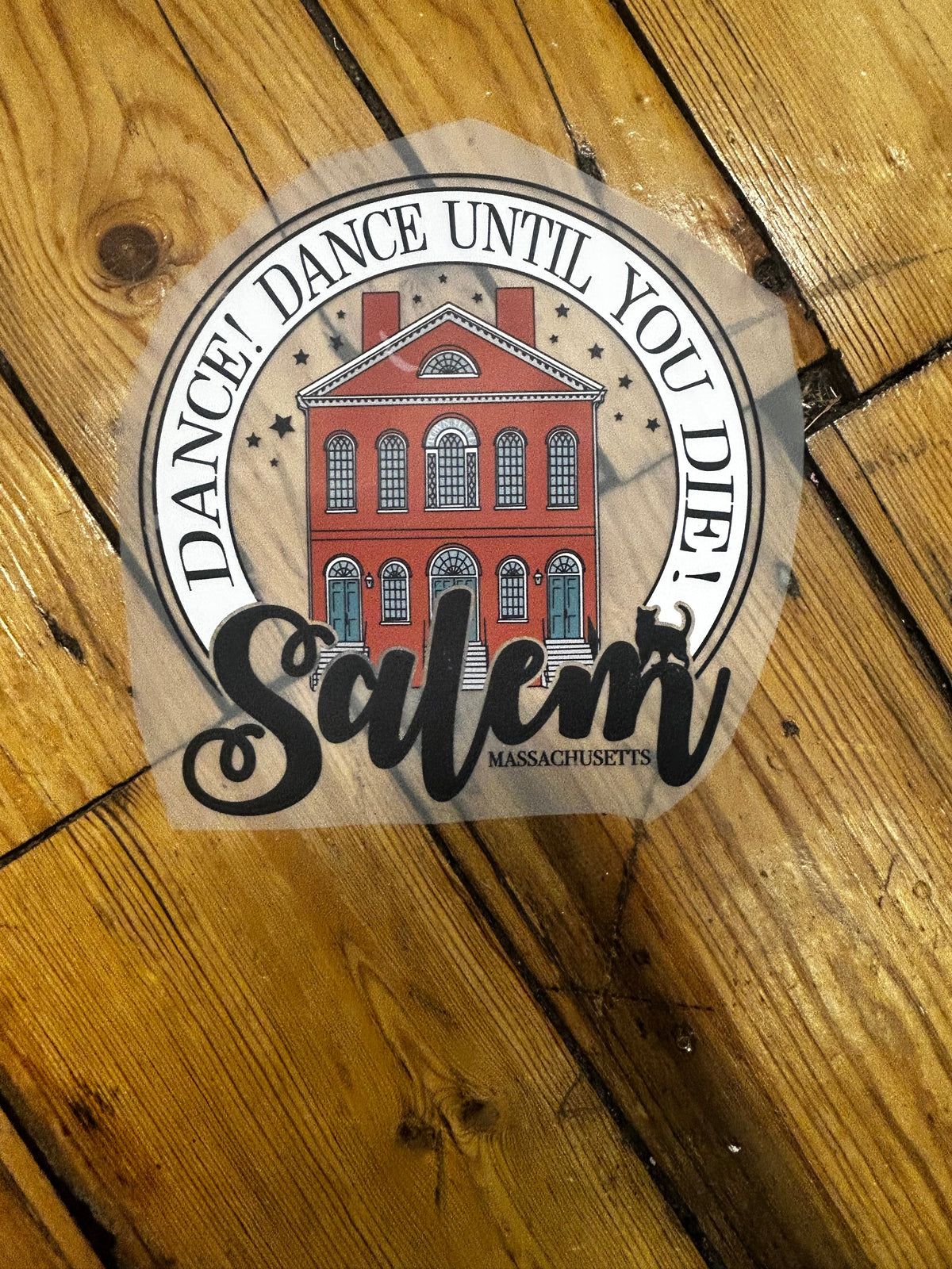 A die-cut sticker featuring a red brick building and the text 'DANCE! DANCE! DANCE! UNTIL YOU DIE!' with a large black script 'Salem' overlay, placed on a wooden floor.