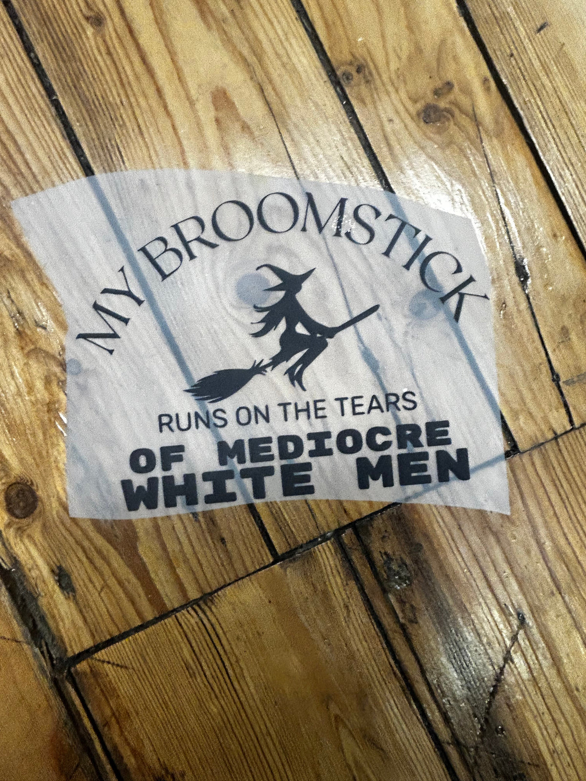 A die-cut vinyl sticker with a black graphic and text reading 'MY BROOMSTICK RUNS ON THE TEARS OF MEDIOCRE WHITE MEN' on a light gray wood-grain background, placed on a wooden floor.