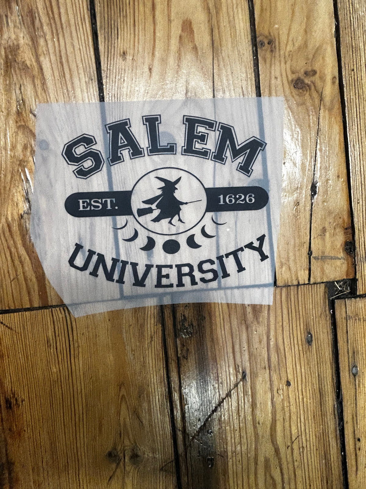 A die-cut vinyl sticker of the Salem University logo, featuring a black graphic print on a translucent white backing, placed on a rustic wooden plank floor.