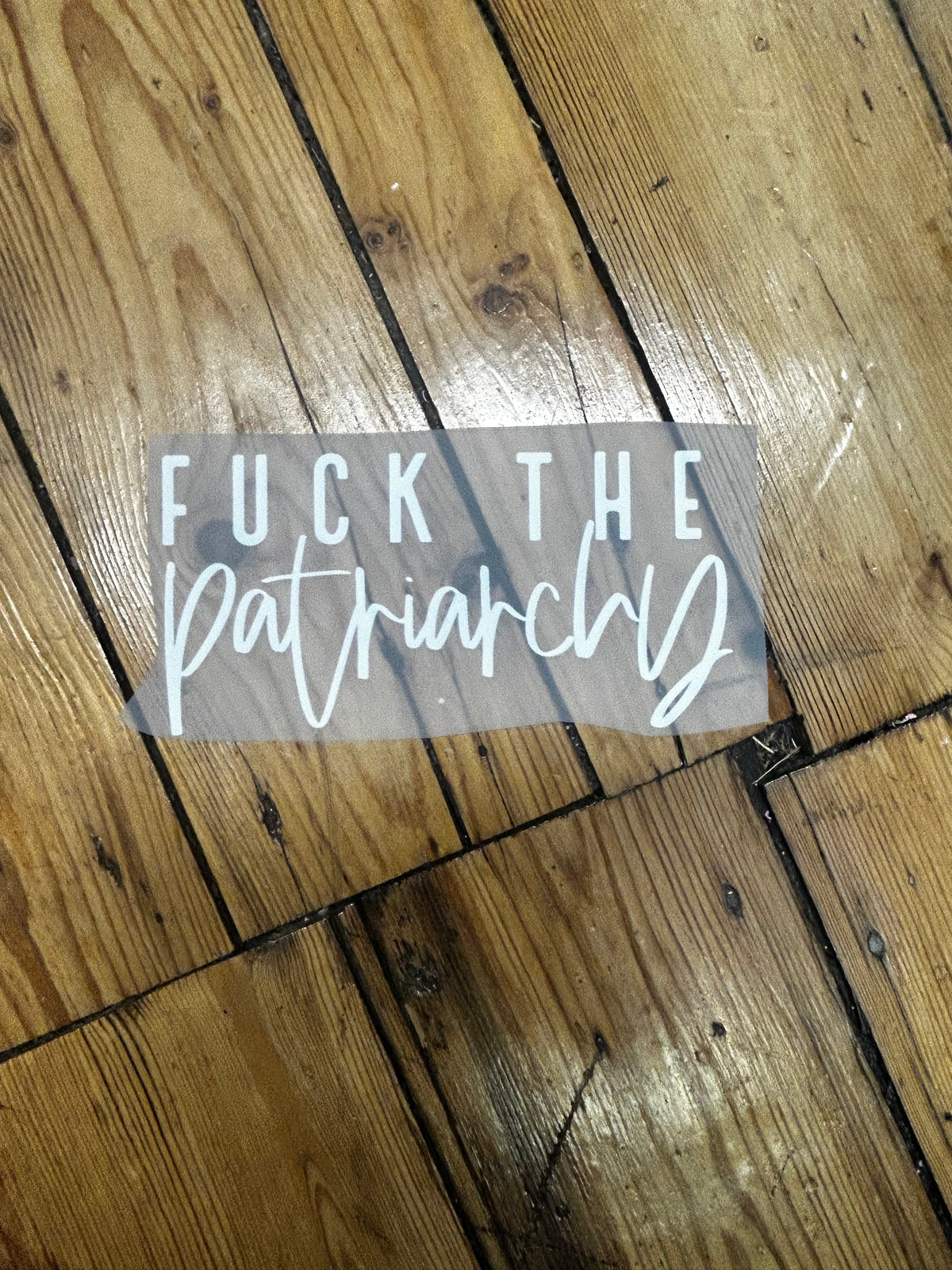 A vinyl decal sticker with the text 'FUCK THE patriarchy' in white, shown partially peeled back from a translucent backing, placed on a rustic wooden plank floor.