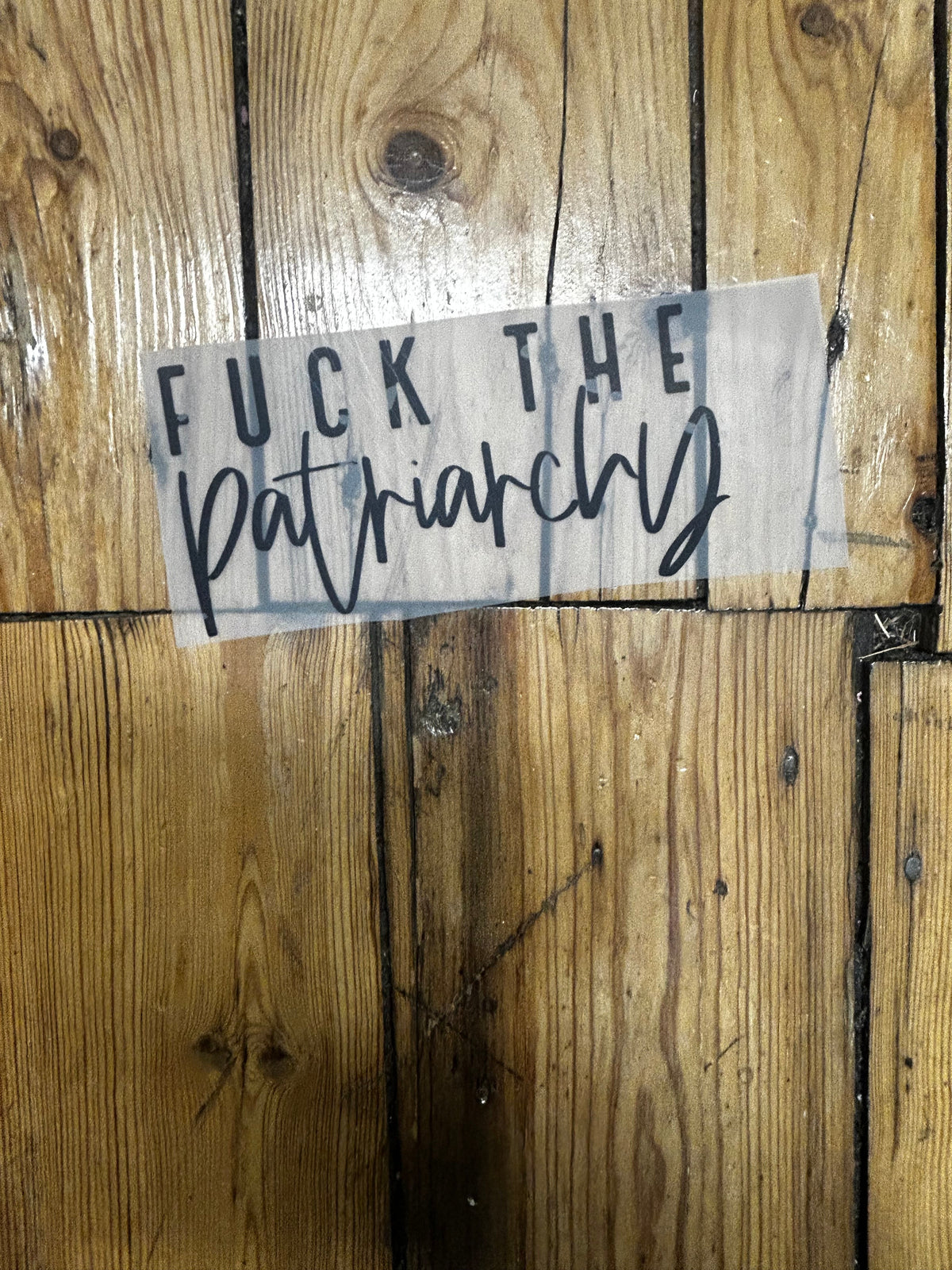 A vinyl decal sticker with the text 'FUCK THE patriarchy' in black script and block lettering, shown applied to a translucent carrier sheet and placed on a rustic wooden plank floor.