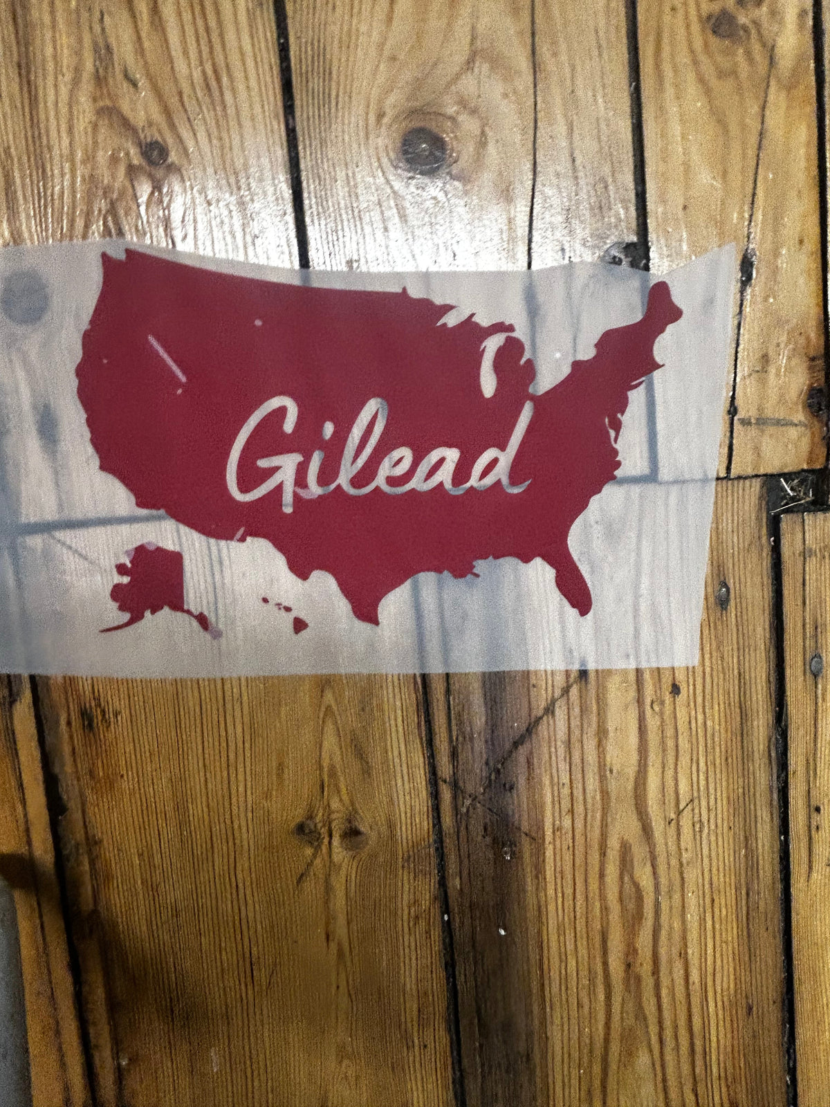 A red and white vinyl decal featuring the outline of the Gilead region with the word 'Gilead' written in white script, shown on a piece of translucent vinyl sheet over a rustic wooden plank background.