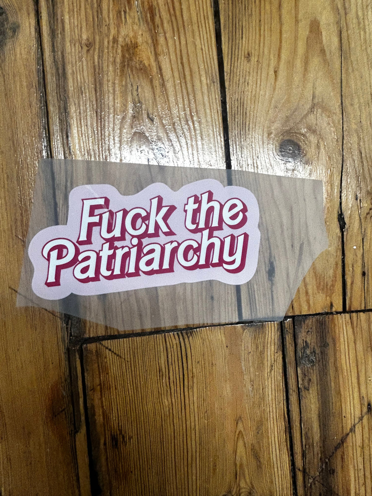 A die-cut vinyl sticker with the text 'Fuck the Patriarchy' in red outlined lettering on a white background, shown on a clear backing sheet over a rustic wooden floor.