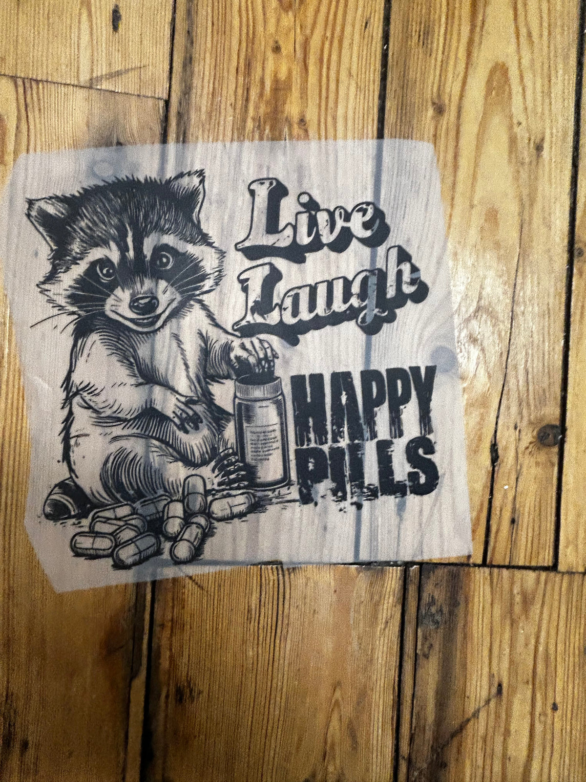 A square white fabric patch or sticker featuring a black graphic of a raccoon holding a pill bottle next to the text 'Live Laugh Pills' and 'HAPPY PILLS', placed on a rustic wooden plank surface.