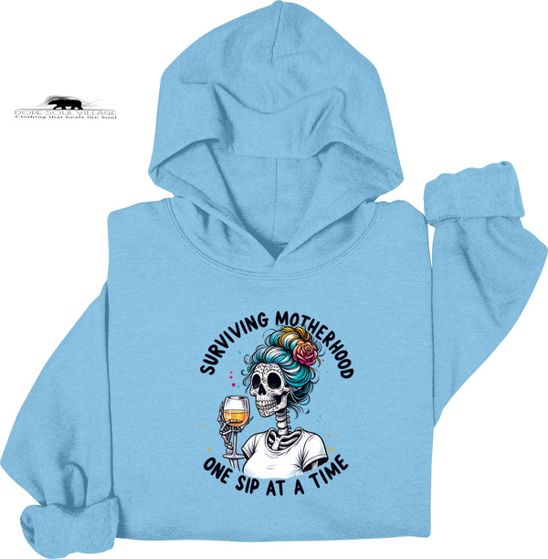 Light blue hoodie with a graphic design featuring a skeleton with colorful hair and a glass of wine, text 'Surviving Motherhood One Sip at a Time', on a white background.