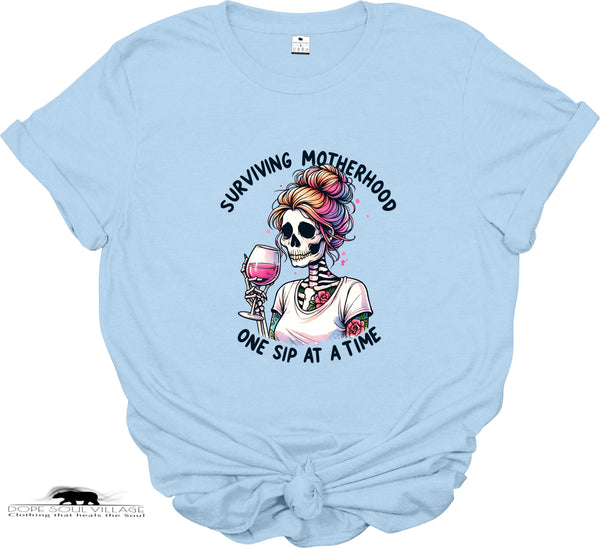 Light blue t-shirt with a graphic of a skeleton holding a glass of wine and text 'Surviving Motherhood One Sip at a Time' on a white background.