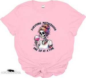 Pink t-shirt with a skeleton holding a glass of wine and text 'Surviving Motherhood One Sip at a Time' on a white background.