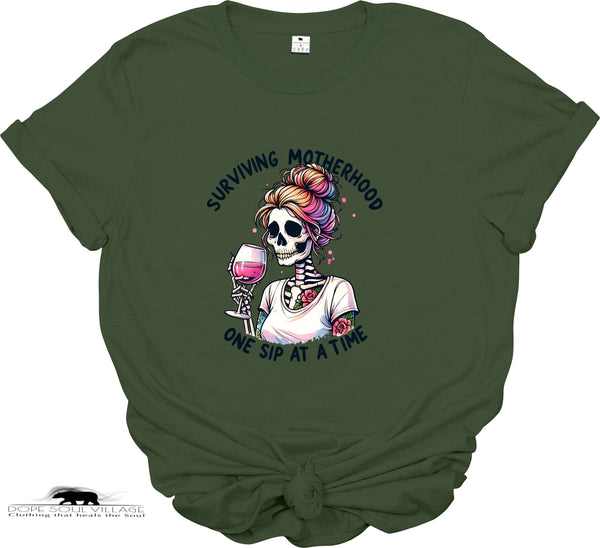 Green t-shirt with a graphic of a skeleton with colorful hair and a glass of wine, text 'Surviving Motherhood One Sip at a Time', on a white background.