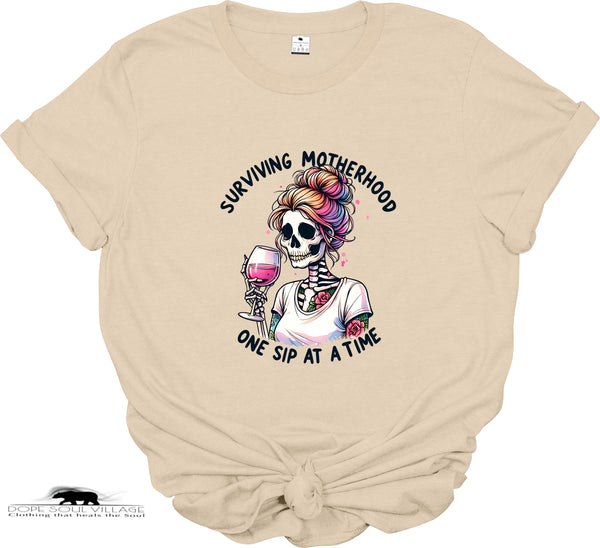 Beige t-shirt with a skeleton holding a glass of wine and text 'Surviving Motherhood One Sip at a Time' on a white background.