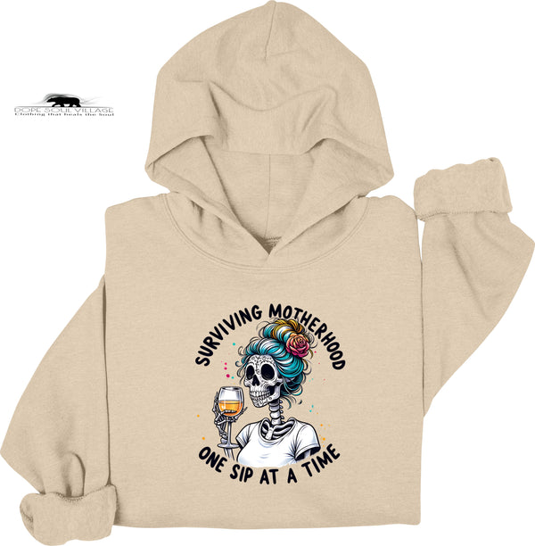 Beige hoodie with a graphic design of a skeleton with colorful hair and a glass of wine, text 'Surviving Motherhood One Sip at a Time', on a white background.