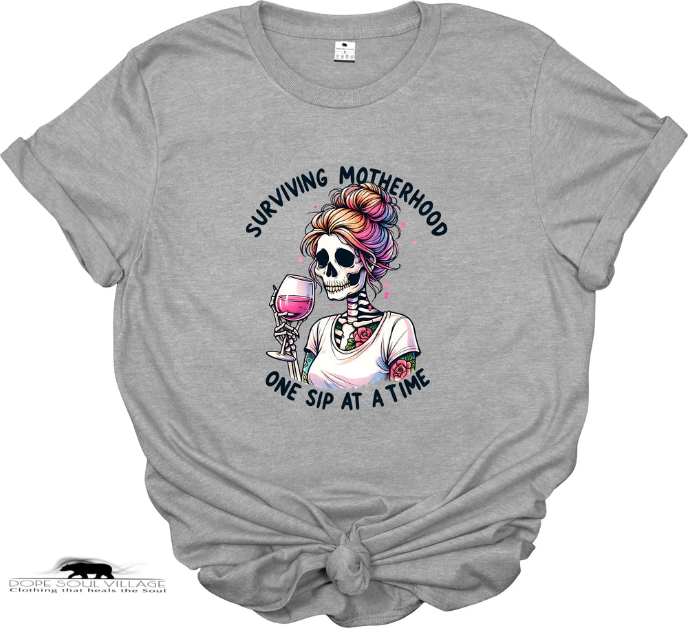 Gray t-shirt with a graphic of a skeleton with colorful hair and text 'Surviving Motherhood One Sip at a Time' on a white background.