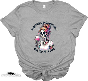 Gray t-shirt with a graphic of a skeleton with colorful hair and text 'Surviving Motherhood One Sip at a Time' on a white background.
