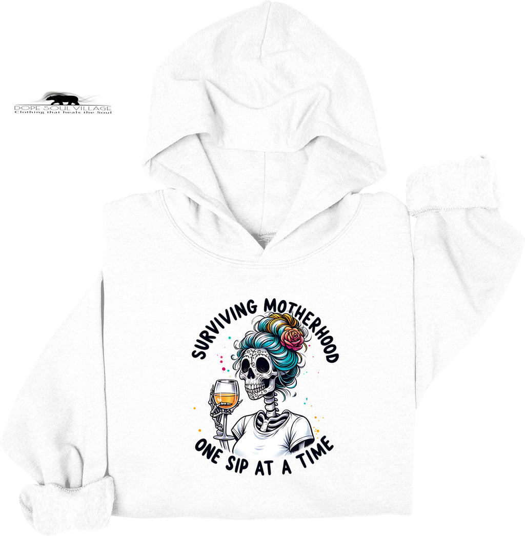 White hoodie with a graphic design of a skeleton with colorful hair holding a glass of wine, text 'Surviving Motherhood One Sip at a Time', and a brand logo on a white background.