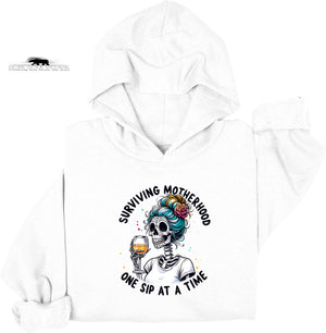 White hoodie with a graphic design of a skeleton with colorful hair holding a glass of wine, text 'Surviving Motherhood One Sip at a Time', and a brand logo on a white background.