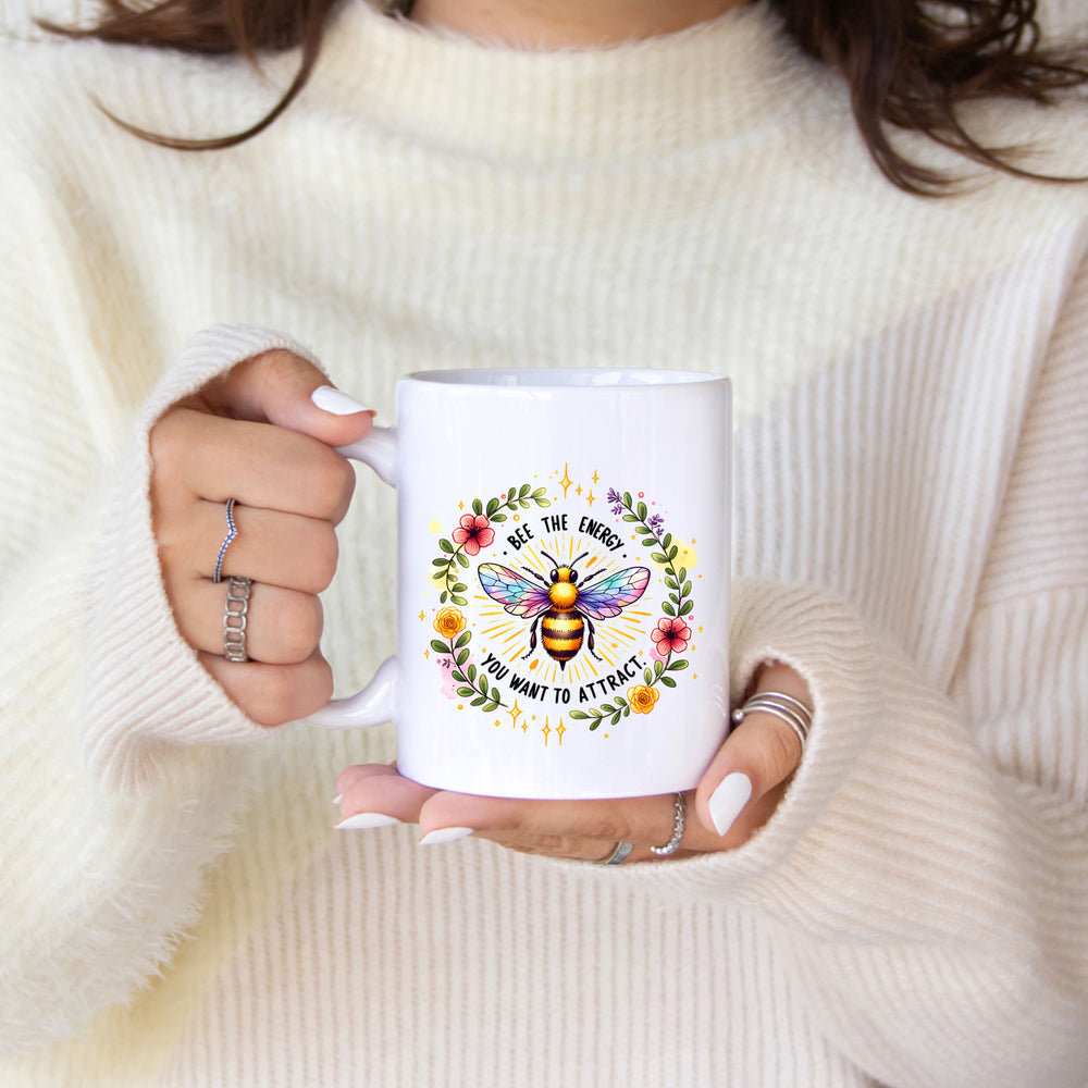 Person holding a white mug with a colorful bee design and text, wearing a cream sweater.