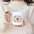 Person holding a white mug with a colorful bee design and text, wearing a cream sweater.