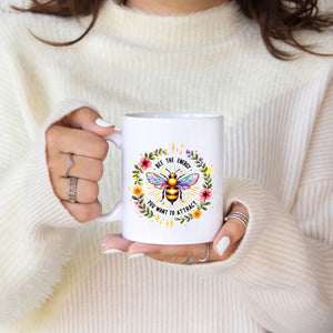 Person holding a white mug with a colorful bee design and text, wearing a cream sweater.