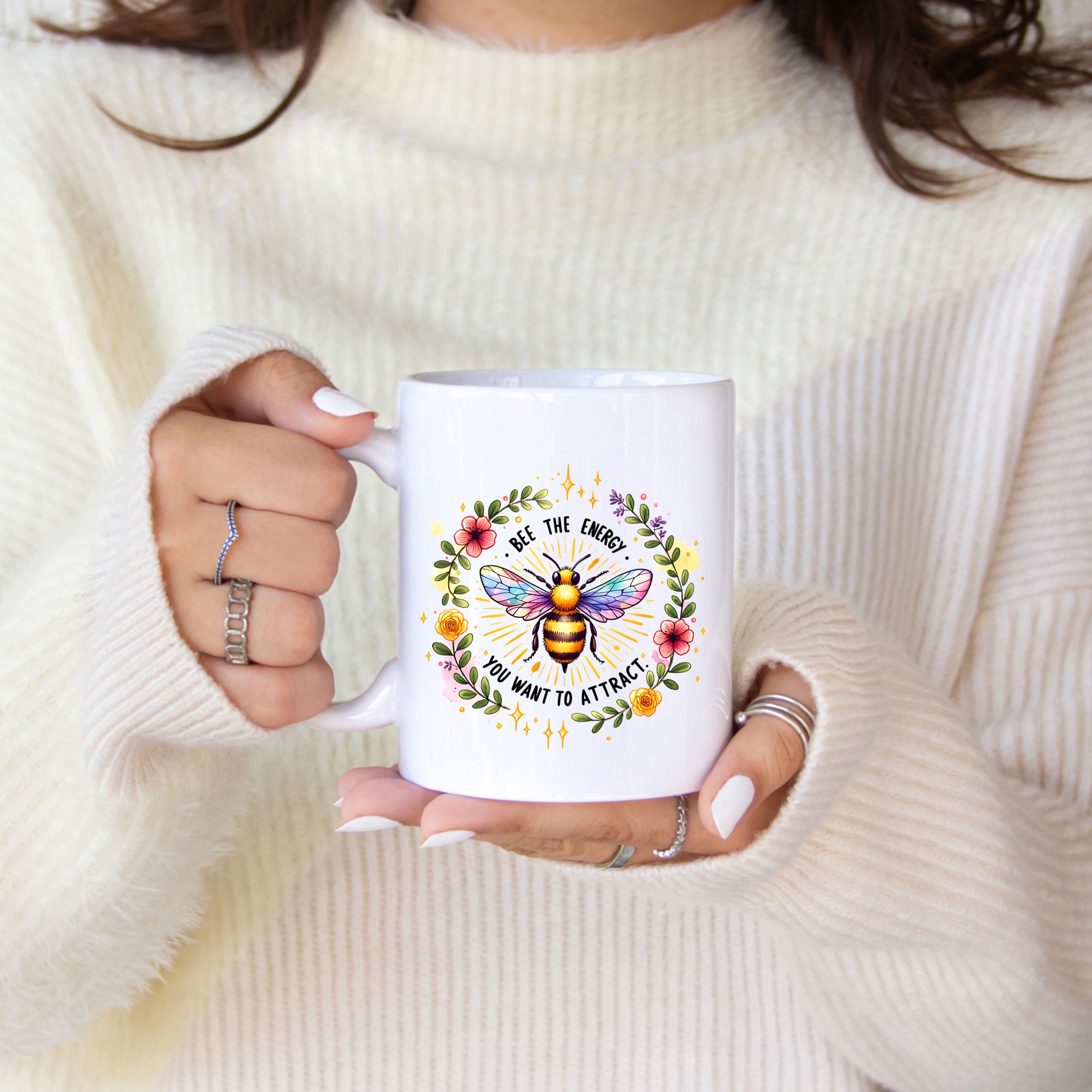 Person holding a white mug with a colorful bee design and text, wearing a cream sweater.