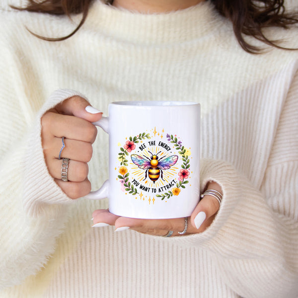 Person holding a white mug with a colorful bee design and text, wearing a cream sweater.