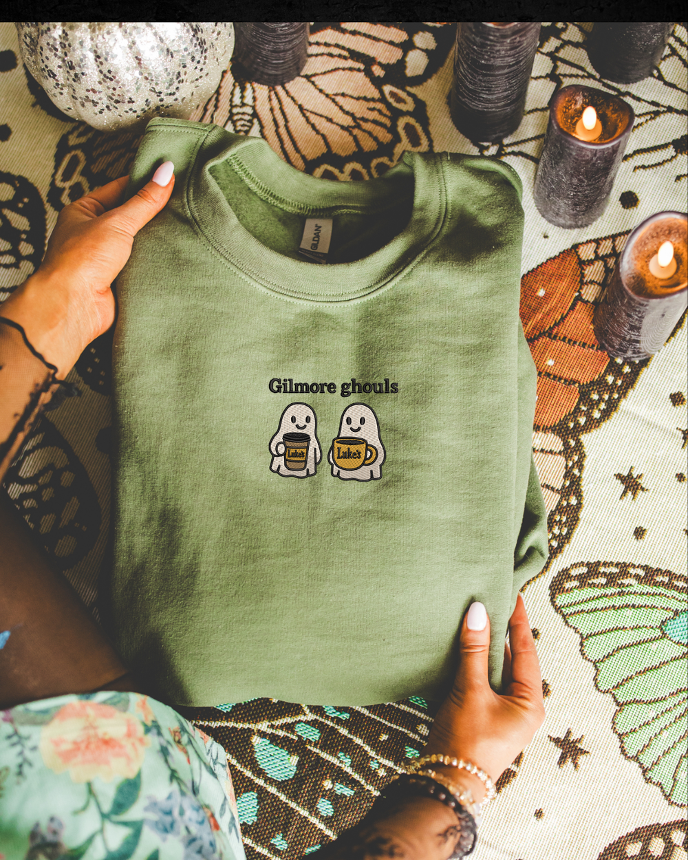 Green t-shirt with 'Gilmore ghosts' design held by a person on a decorative surface with candles.