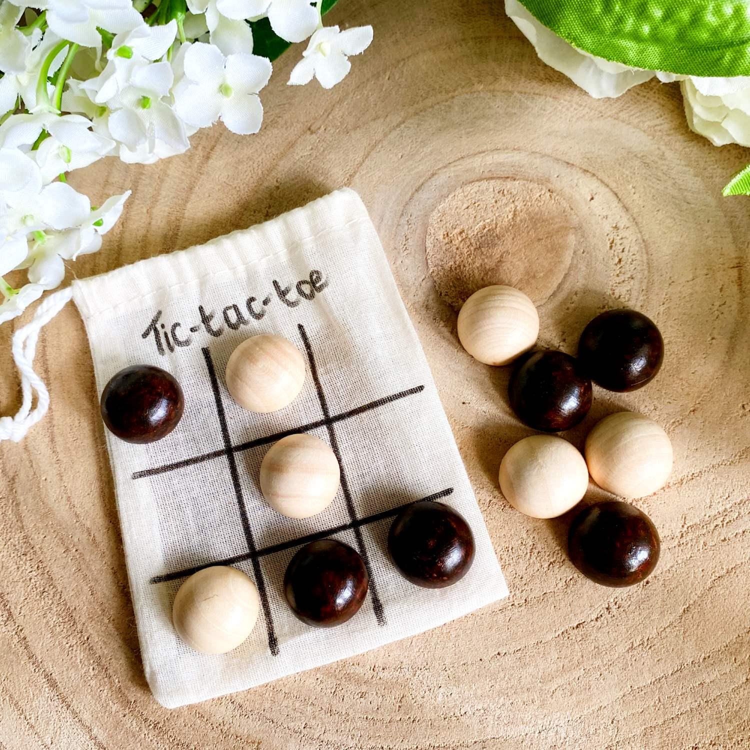 Wooden tic-tac-toe (noughts and crosses) game with its cotton storage bag lying on a wooden table with white flowers in the background.