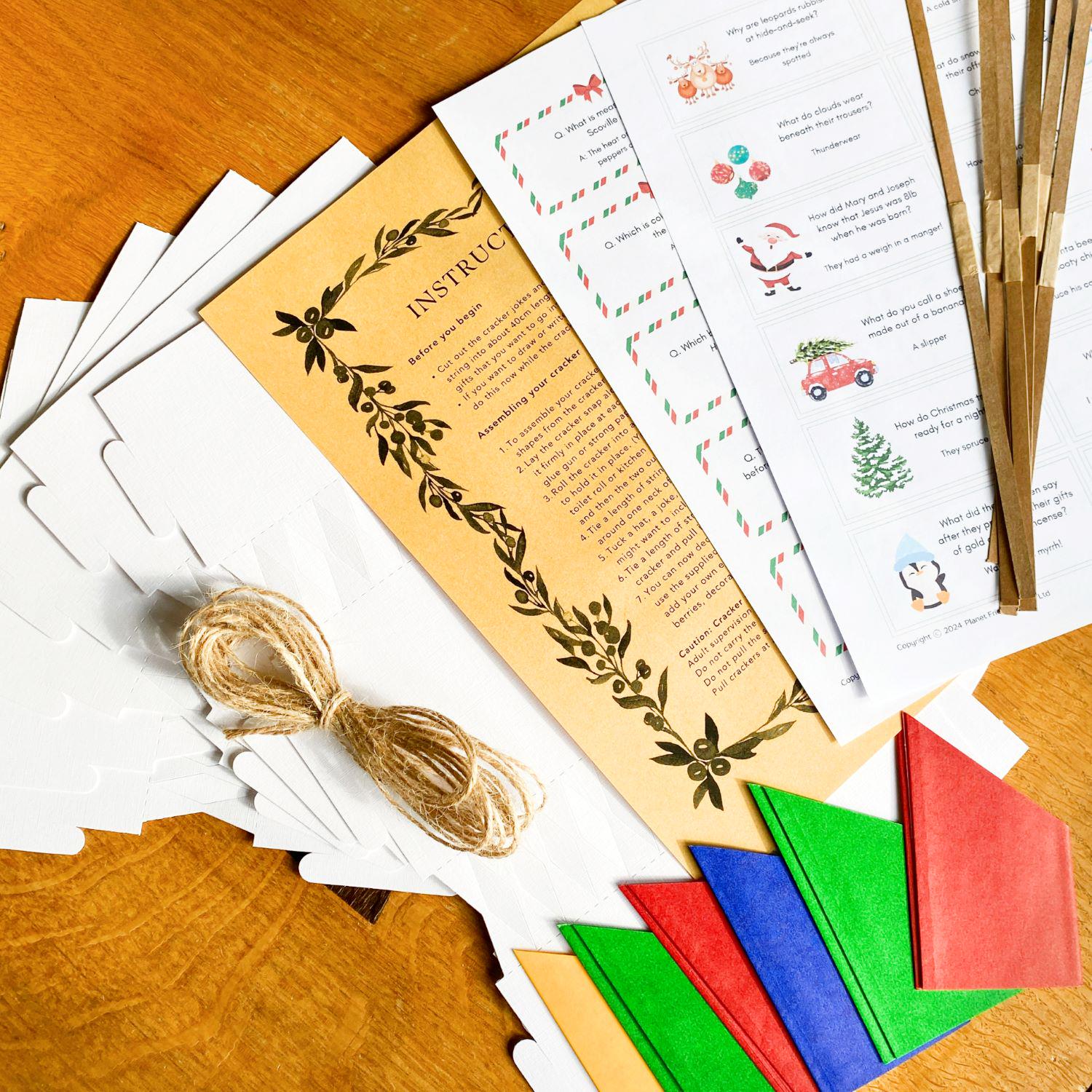Contents of the white version of the Eco-Friendly Christmas Cracker Kit spread out on a wooden table to show: white cracker blanks, multi-coloured tissue paper hats, twine, cracker snaps, printed instructions, sheet of cracker jokes and sheet of trivia questions.