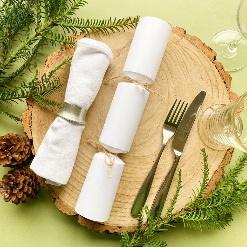 A homemade white Christmas cracker resting on a Christmas place-setting with white napkin, glasses and cutlery alongside