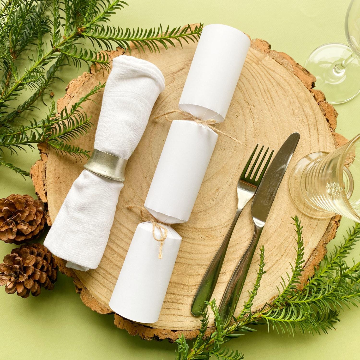 A homemade white Christmas cracker resting on a Christmas place-setting with white napkin, glasses and cutlery alongside